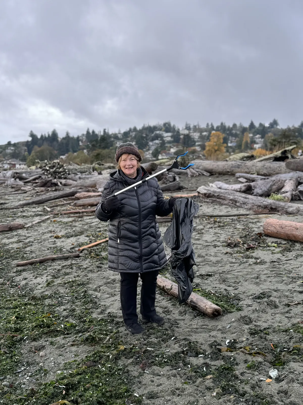 A member in a pink jacket holding a garbage bag on a driftwood beach